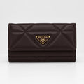 A dark brown Prada quilted leather wallet with a gold-tone triangular logo plaque on the front, set against a plain light gray background.