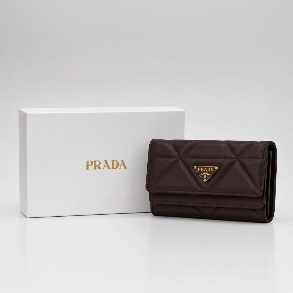 A dark brown Prada quilted wallet with its signature gold Tri-Fur logo plaque, displayed next to its white presentation box with gold Prada branding on a clean white background.