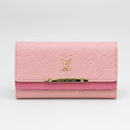 A pink Louis Vuitton monogram wallet with a gold-tone metal bar engraved with 'LOUIS VUITTON' and an embossed LV logo on the front flap, set against a plain light gray background.